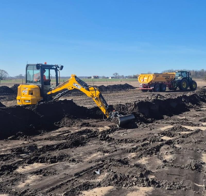 Minikoparka JCB przy wykopie, za nią ciągnik z przyczepą budowlaną